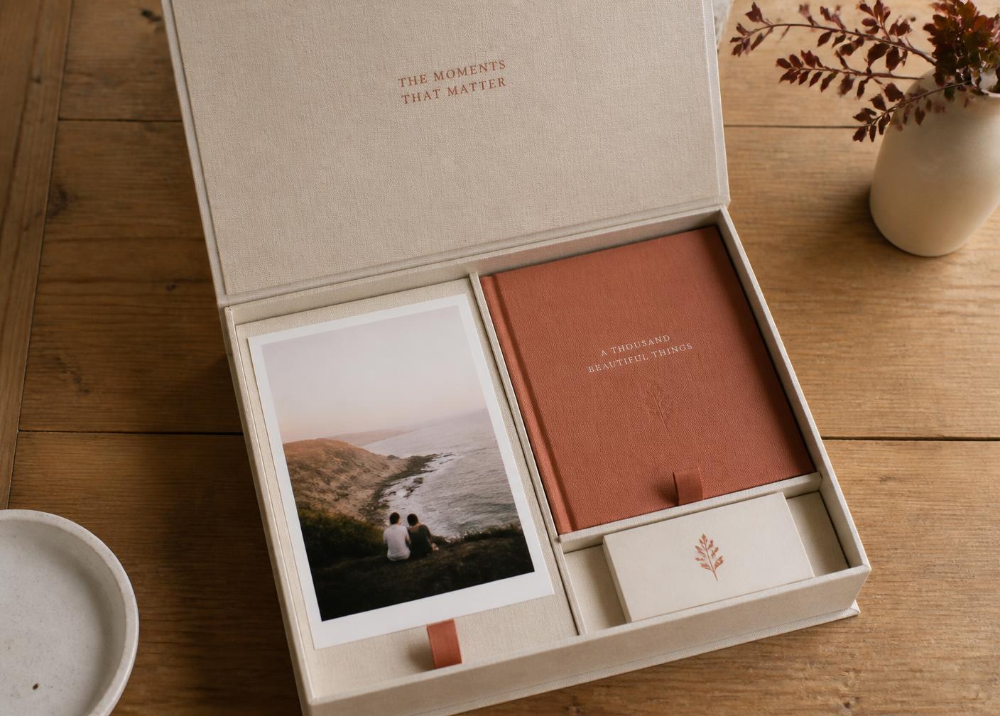 An open linen keepsake box holding printed photographs and a hardcover book